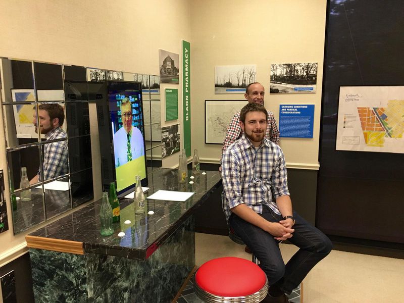 Jeremy Murray, rear, and Kyle Mathers, respectively the Kenilworth Historical Society’s collections manager and curator, sit at the Blann Pharmacy soda fountain mockup central to “How Can I Help You: Business and Commerce in Kenilworth.” The mockup allows visitors to hear an oral history of the pharmacy, from Robert Stensby, its last owner. The exhibition runs through October.(Kathy Routliffe/Pioneer Press)